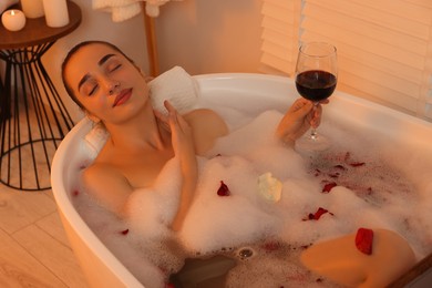 Photo of Woman with glass of wine taking bath in tub with foam and rose petals indoors
