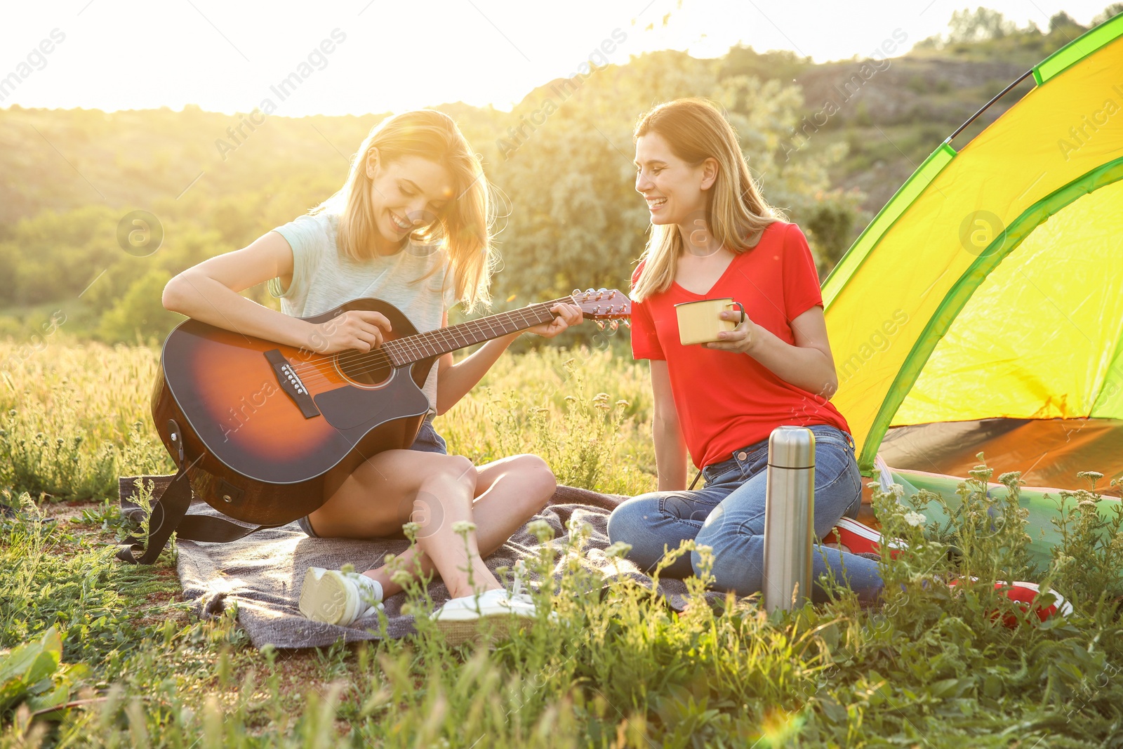 Photo of Young women resting with hot drink and guitar near camping tent in wilderness