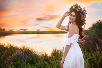 Photo of Young woman wearing wreath made of beautiful flowers outdoors at sunset