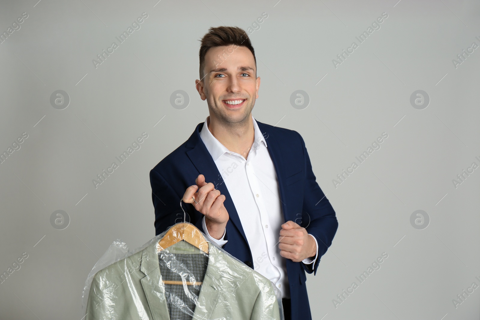 Photo of Man holding hanger with jacket in plastic bag on light grey background. Dry-cleaning service