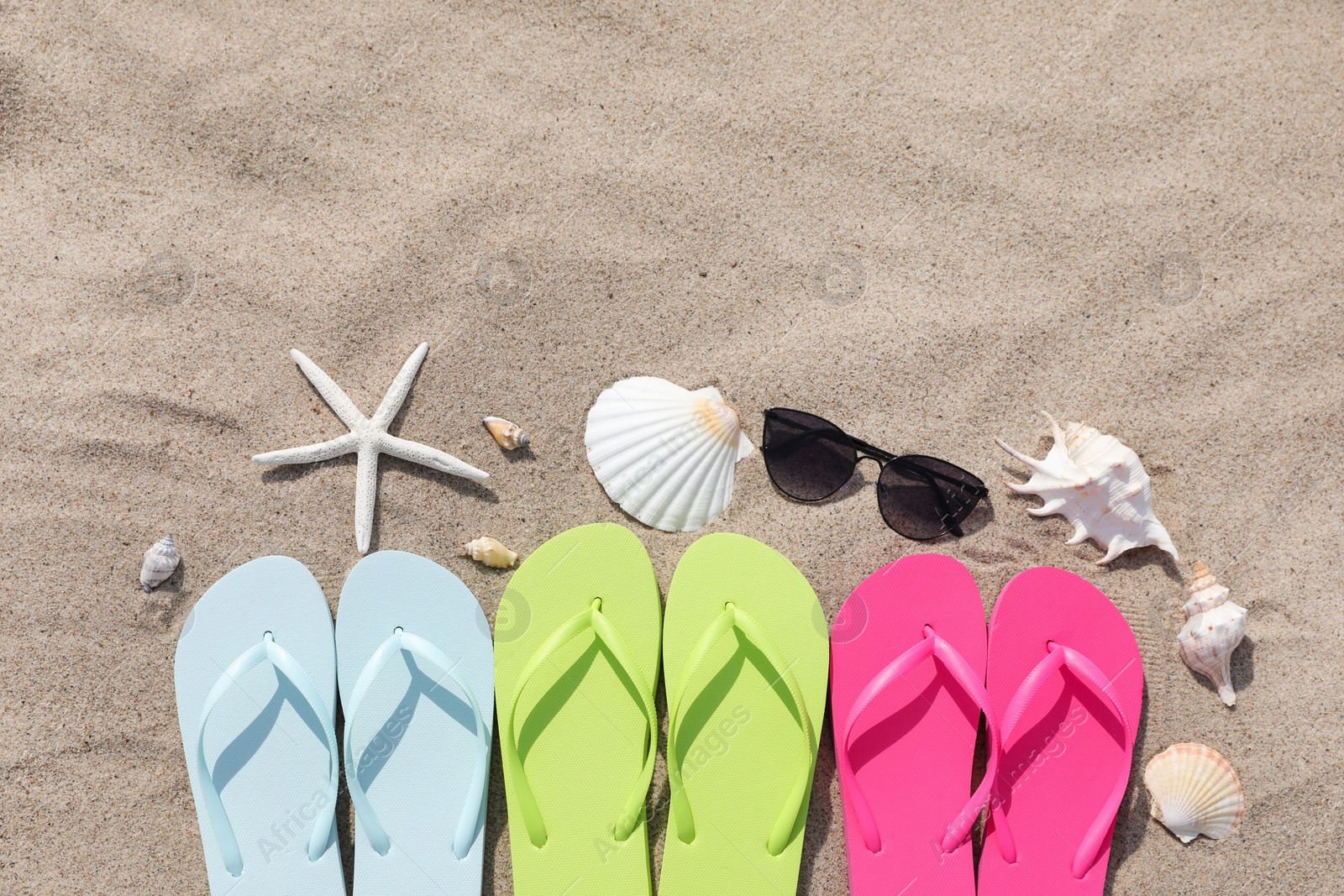 Photo of Stylish colorful flip flops, sunglasses, seashells and starfish on sand outdoors, flat lay. Space for text