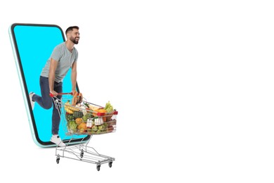 Grocery shopping via internet. Happy man with shopping cart full of products running out of huge smartphone on white background