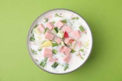 Delicious cold summer soup (okroshka) with boiled sausage in bowl on green table, top view