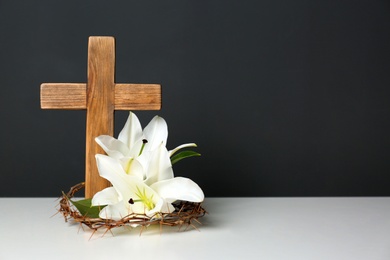 Photo of Wooden cross, crown of thorns and blossom lilies on table against color background, space for text