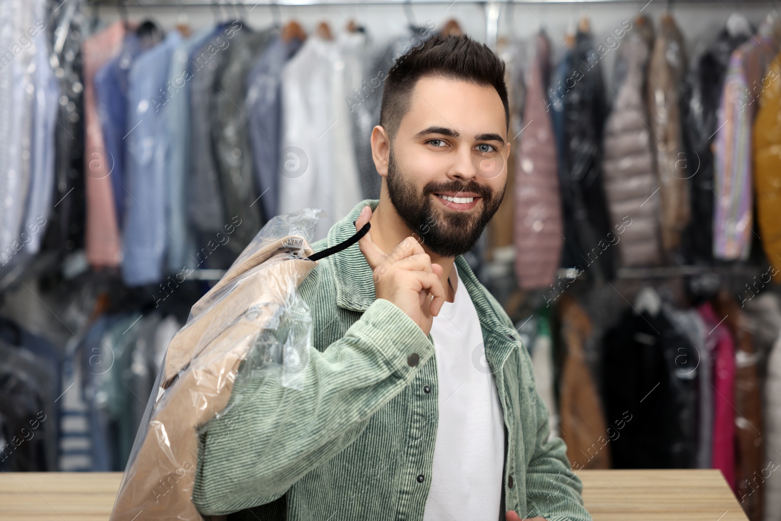 Photo of Dry-cleaning service. Happy man holding hanger with coat in plastic bag indoors