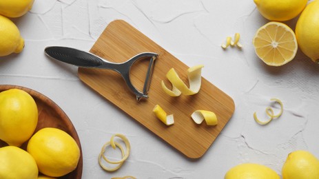 Photo of Wooden board, lemons, peeler and fresh rind on white textured table, flat lay