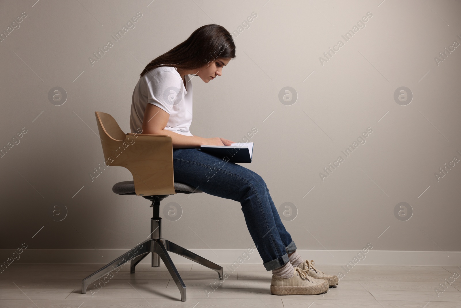 Photo of Young woman with bad posture reading book while sitting on chair near grey wall. Space for text
