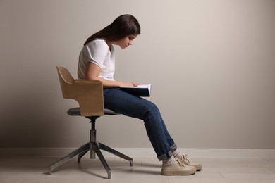 Photo of Young woman with bad posture reading book while sitting on chair near grey wall. Space for text