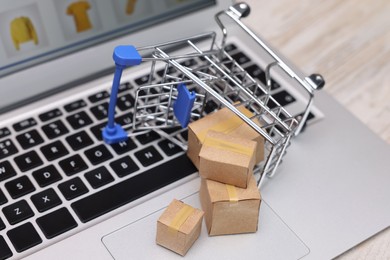 Photo of Internet store. Small cardboard boxes, shopping cart and laptop on light wooden table, closeup