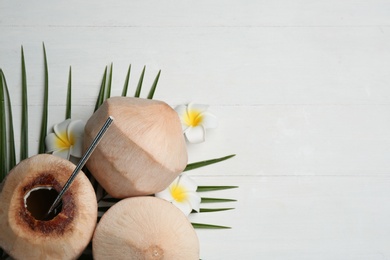 Photo of Young peeled coconuts with straw, tropical leaf and flowers on white wooden table, flat lay. Space for text