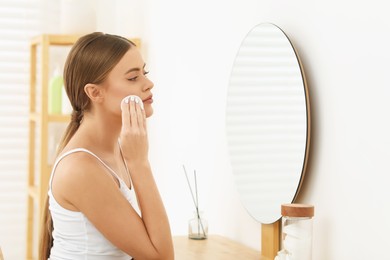 Beautiful woman removing makeup with cotton pad near mirror indoors