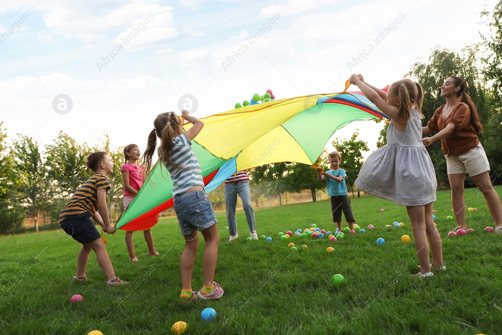 Photo of Group of children and teacher playing with rainbow playground parachute on green grass. Summer camp activity