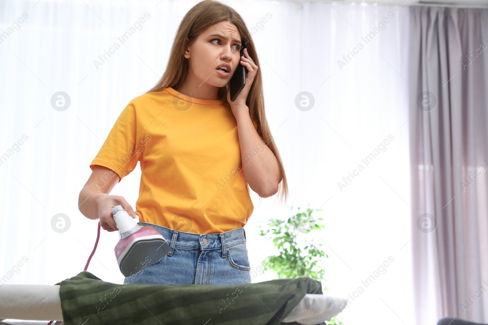 Photo of Emotional woman talking on phone while ironing clothes at home. Space for text