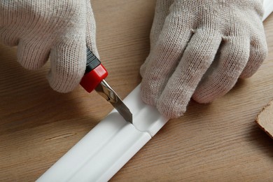Photo of Worker cutting foam crown molding with utility knife at wooden table, closeup
