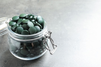 Photo of Glass jar with green spirulina pills on grey table, space for text