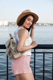 Beautiful young woman with stylish backpack and hat near river