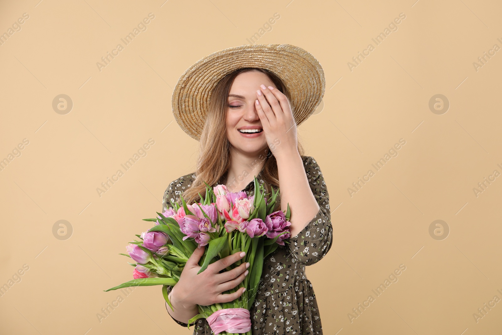 Photo of Happy young woman in straw hat holding bouquet of beautiful tulips on beige background