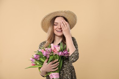 Happy young woman in straw hat holding bouquet of beautiful tulips on beige background