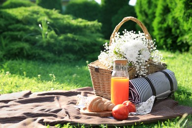 Photo of Picnic blanket with delicious food, juice, rolled mat, flowers and wicker basket in garden on sunny day