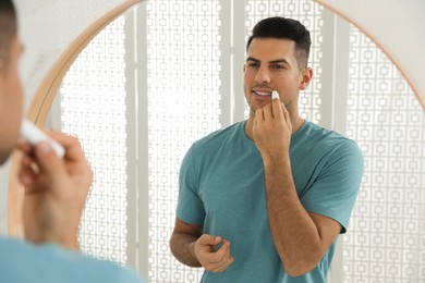 Photo of Man applying hygienic lip balm in front of mirror at home