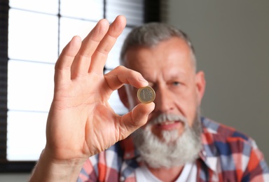 Senior man holding coin indoors, focus on hand. Space for text