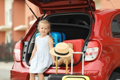 Little girl sitting in car trunk with suitcases outdoors