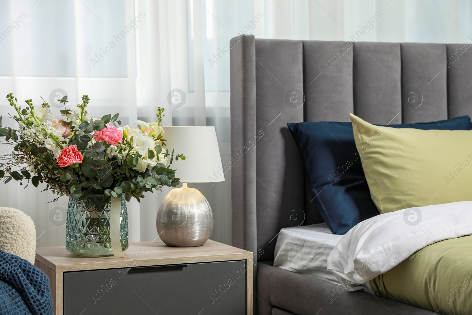 Photo of Lamp and beautiful bouquet on wooden bedside table near comfortable bed in room. Stylish interior