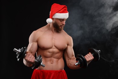 Muscular young man in Santa hat with dumbbells on black background