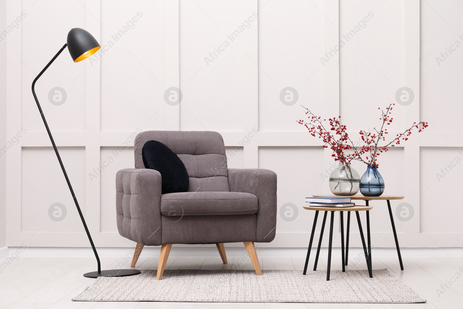 Photo of Hawthorn branches with red berries in vases, armchair and lamp indoors. Interior design
