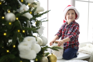 Photo of Cute little boy in Santa hat holding Christmas candleholder on window sill at home