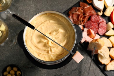 Fork with piece of ham, fondue pot with melted cheese and snacks on grey table, flat lay