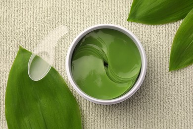 Photo of Jar of under eye patches with spoon and green leaves on light cloth, flat lay. Cosmetic product