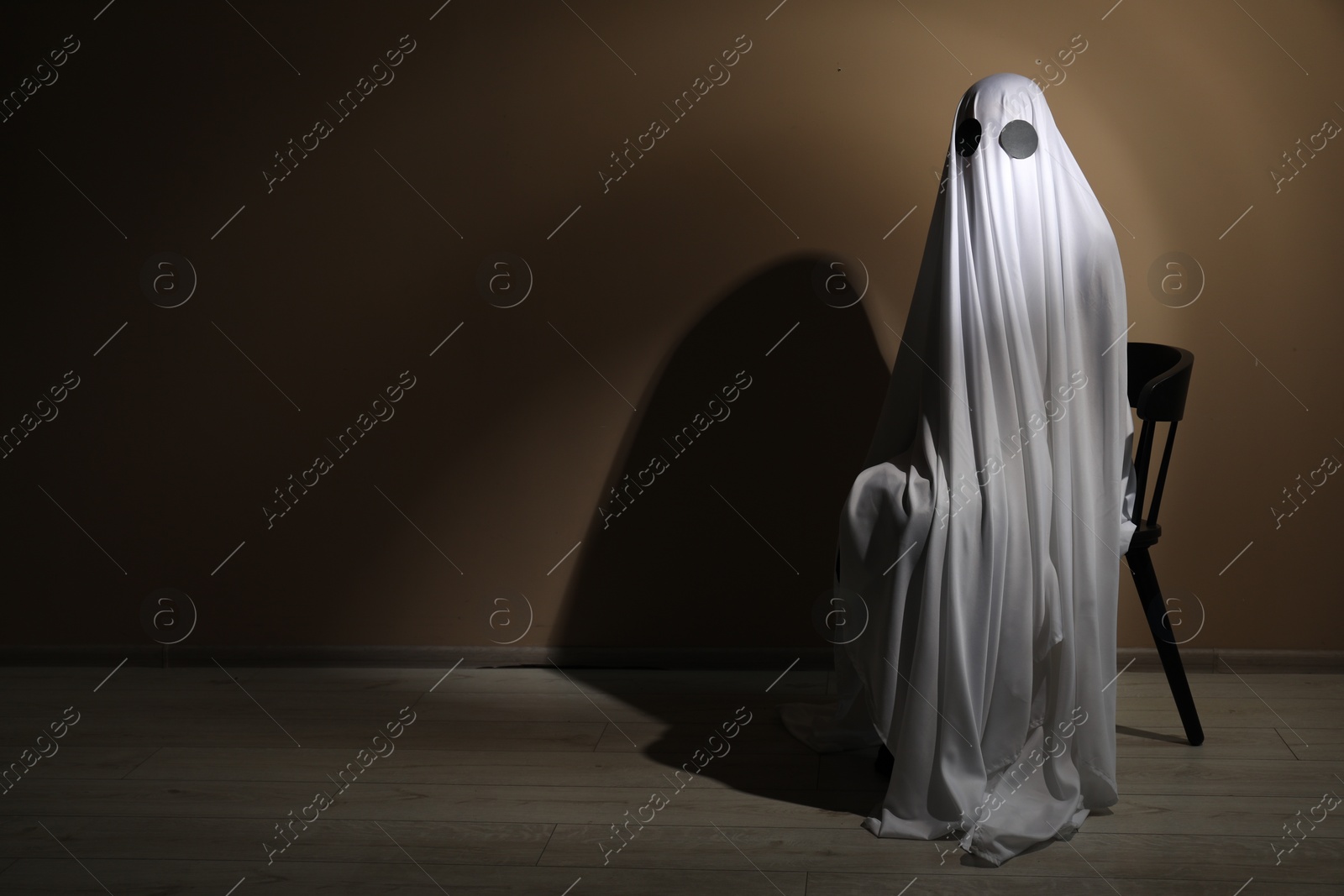 Photo of Creepy ghost. Woman covered with sheet sitting on chair near brown wall, space for text