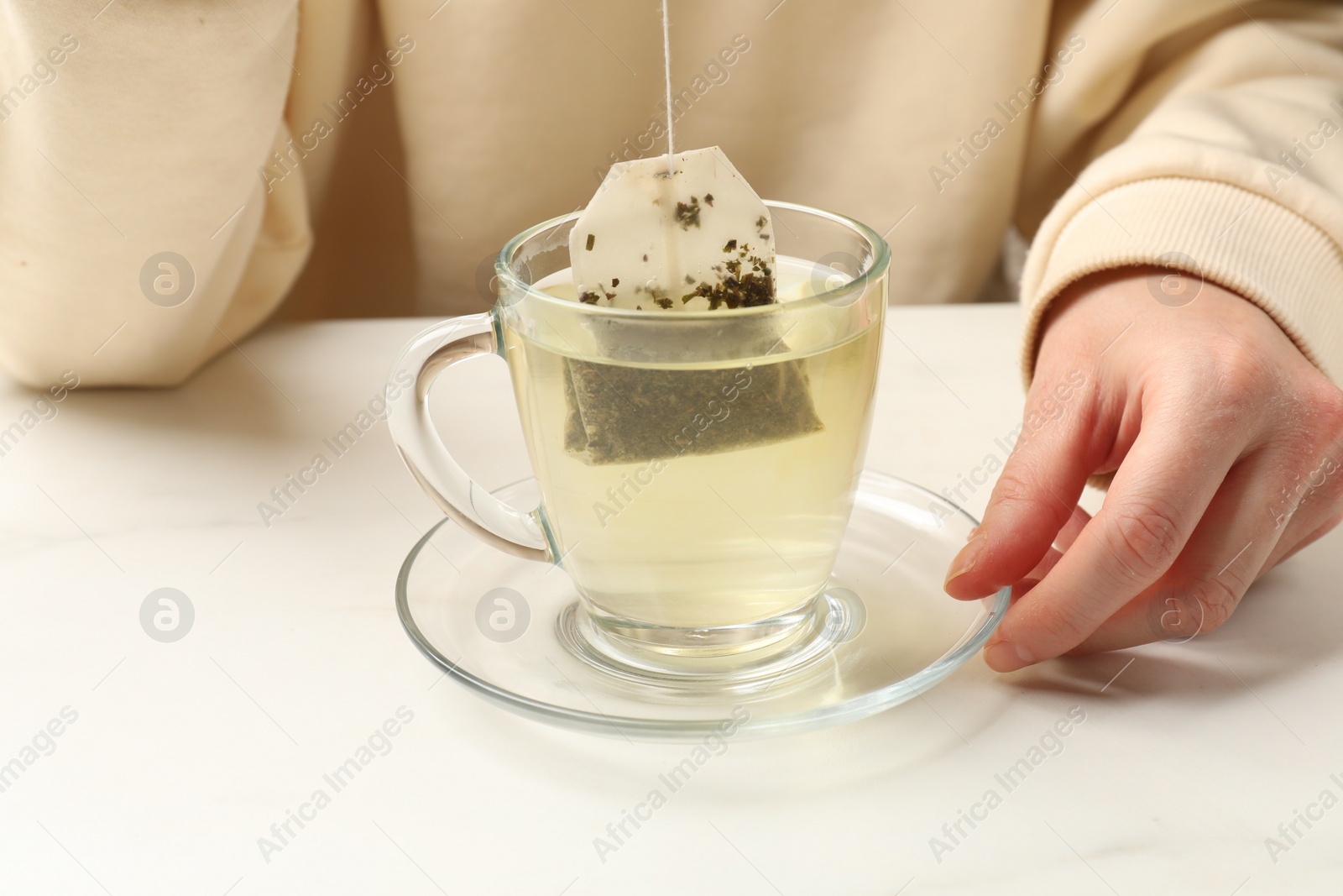 Photo of Woman putting tea bag in glass cup with hot water at white table, closeup