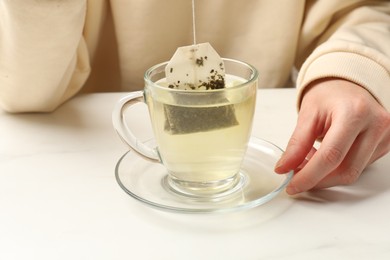 Photo of Woman putting tea bag in glass cup with hot water at white table, closeup