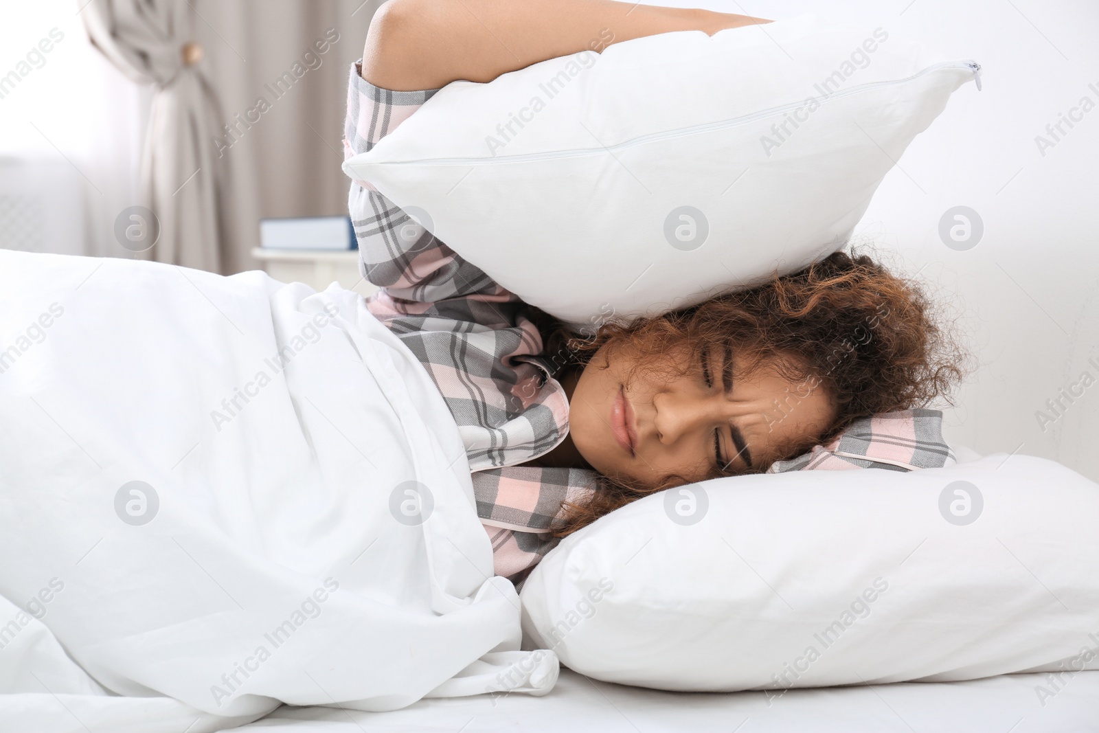 Photo of Young African-American woman covering ears with pillows at home. Bedtime