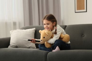 Photo of Little girl changing TV channels with remote control on sofa at home