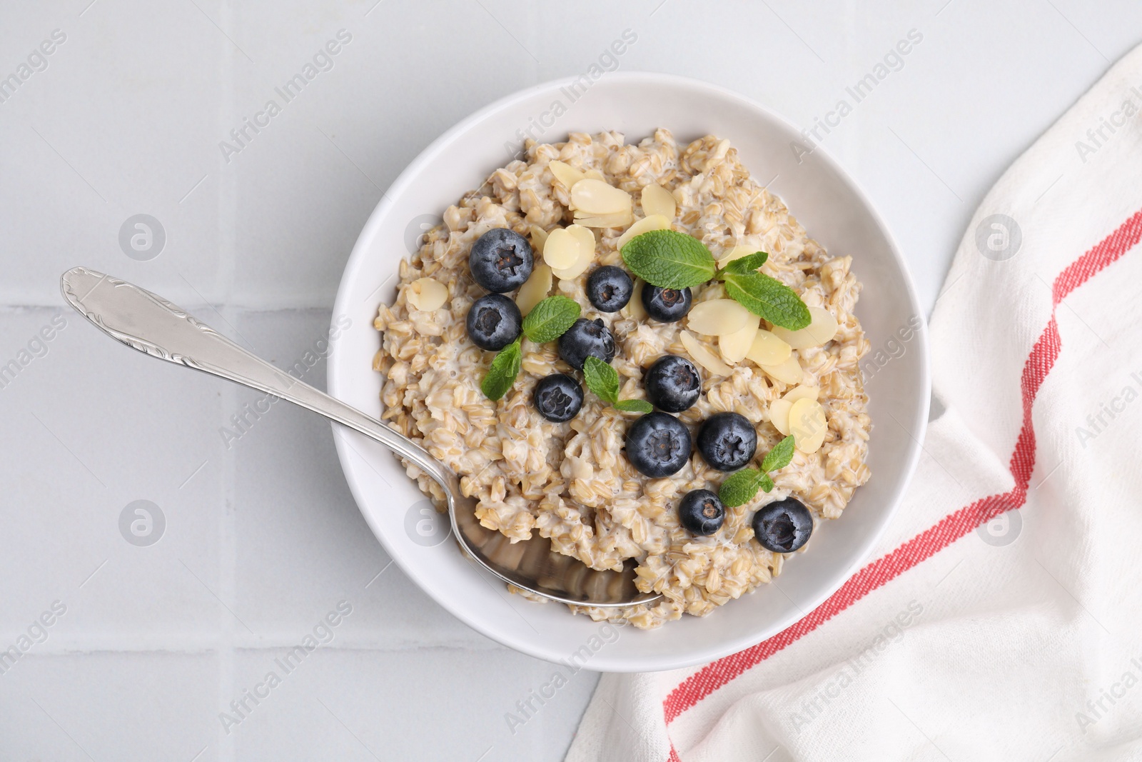 Photo of Tasty oatmeal with blueberries, mint and almond petals in bowl on white tiled table, top view