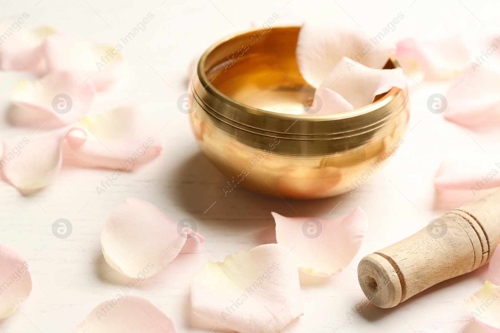 Photo of Golden singing bowl with petals and mallet on white wooden table, closeup. Sound healing