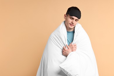Man in pyjama and sleep mask wrapped in blanket on beige background, space for text