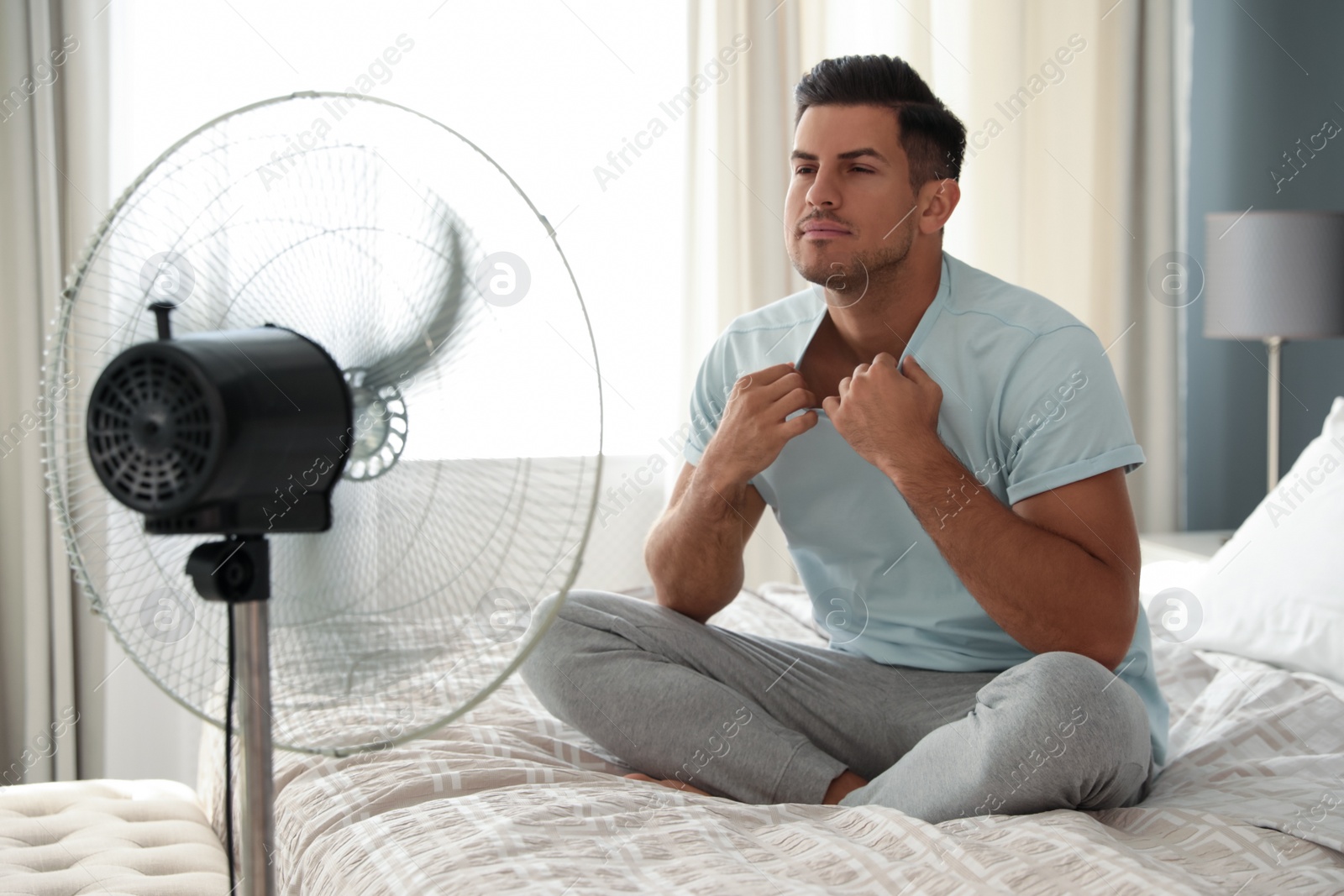 Photo of Man enjoying air flow from fan on bed in room. Summer heat