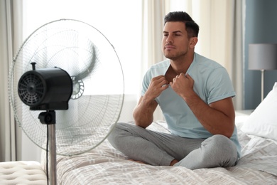 Photo of Man enjoying air flow from fan on bed in room. Summer heat