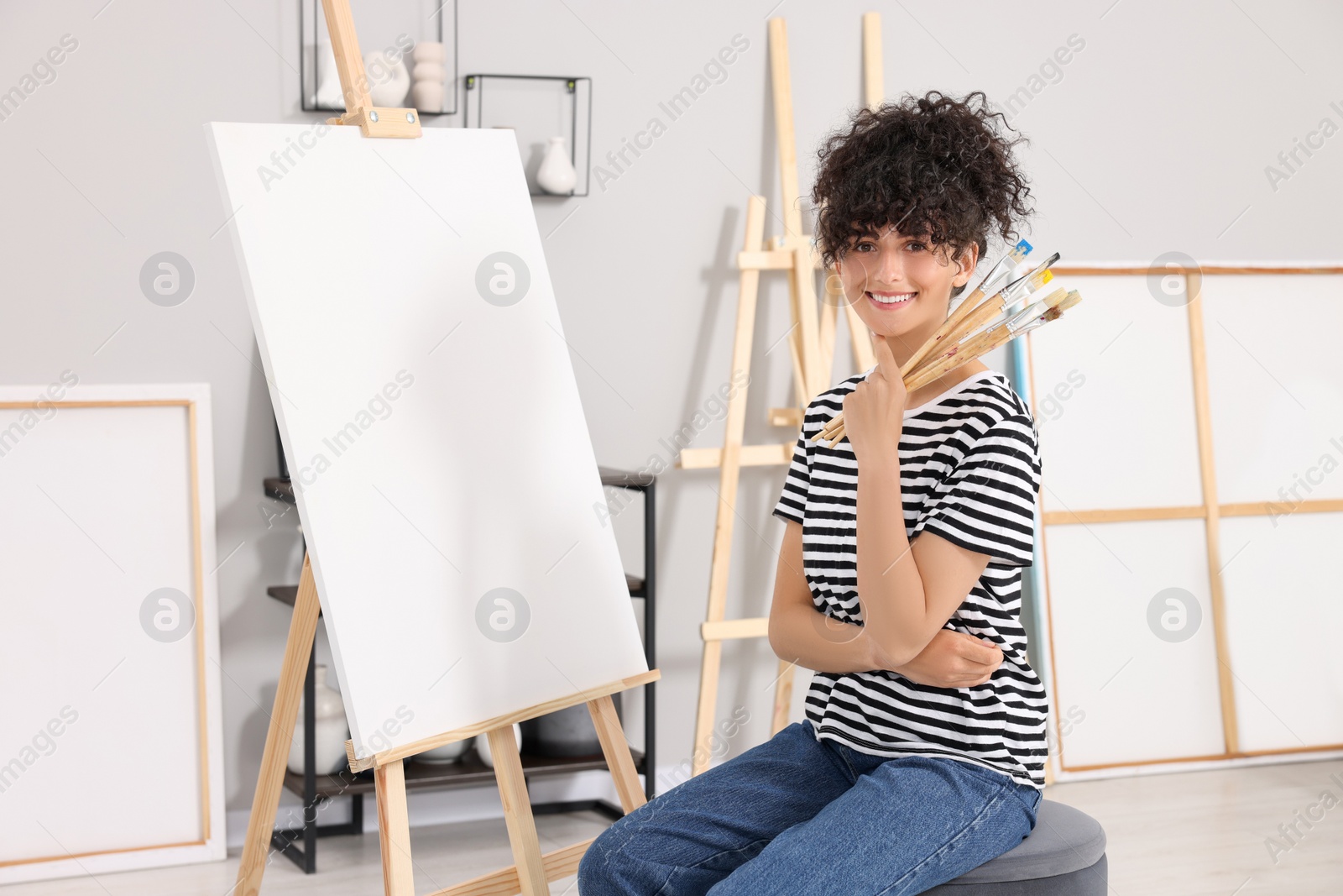 Photo of Young woman holding brushes near easel with canvas in studio