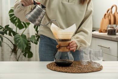 Photo of Woman pouring hot water into glass chemex coffeemaker with paper filter and coffee at white table, closeup