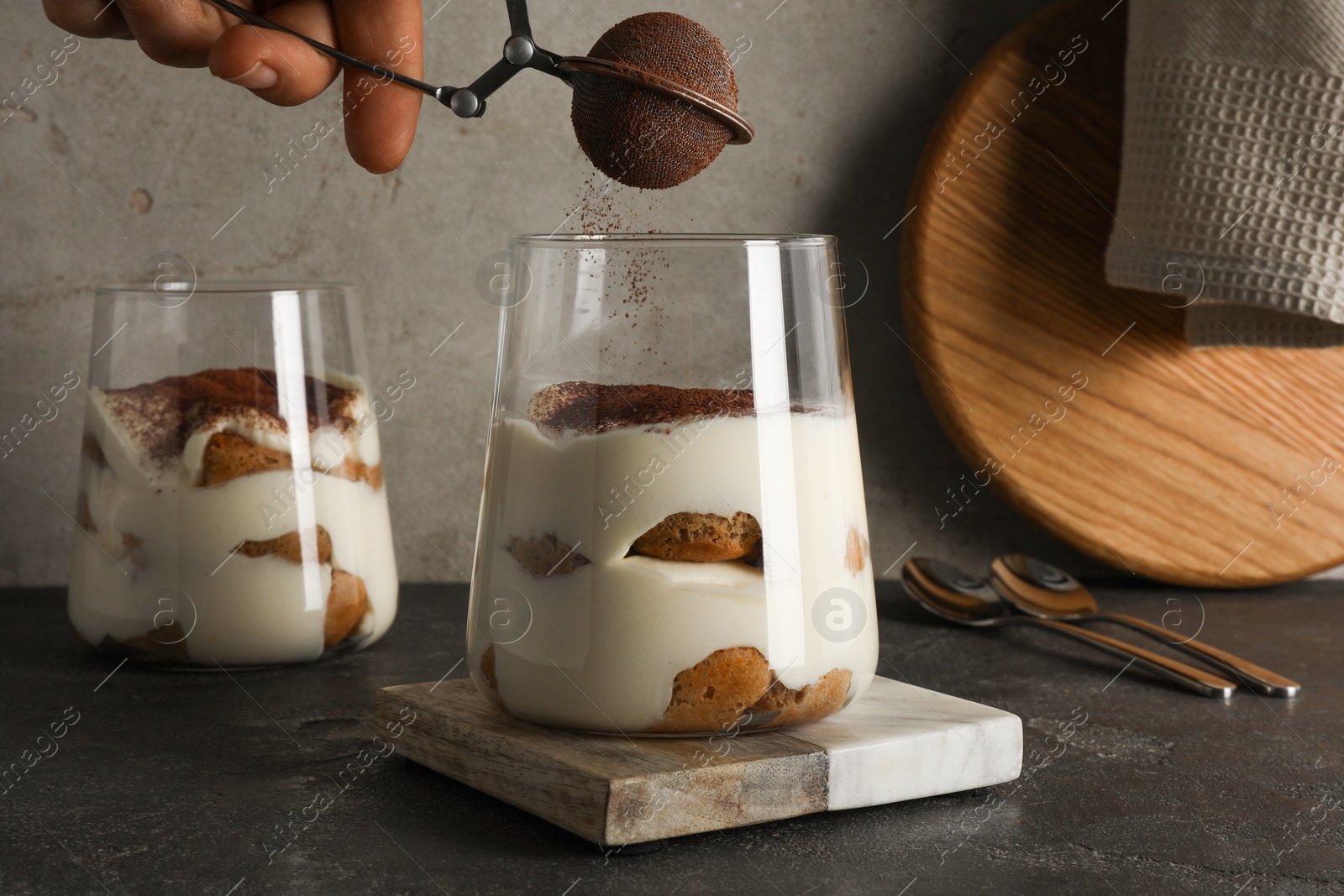 Photo of Woman dusting delicious tiramisu with cocoa powder on grey table, closeup