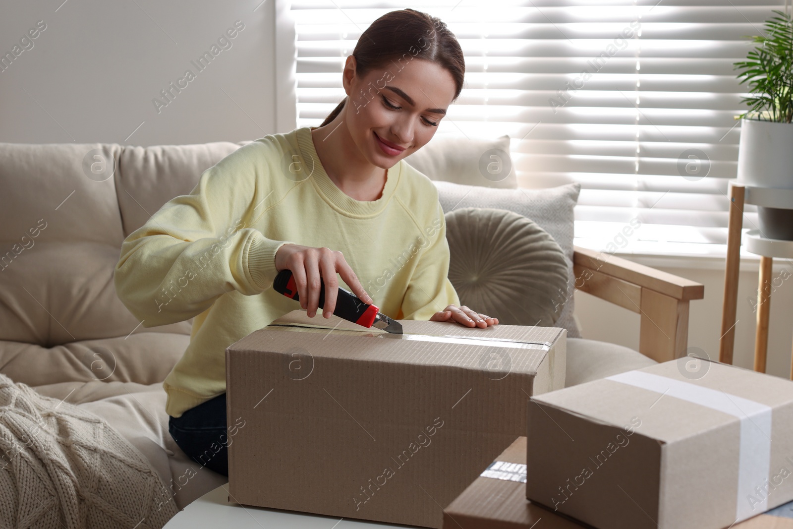 Photo of Young woman using utility knife to open parcel at home