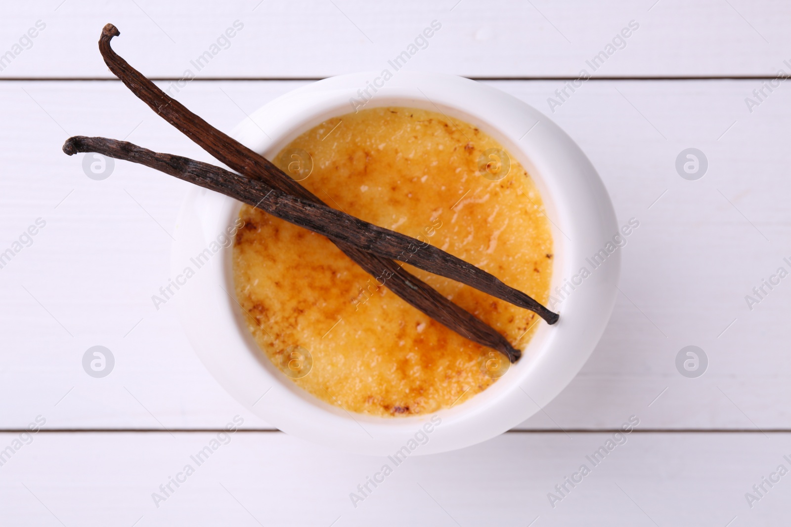Photo of Delicious creme brulee in bowl and vanilla pods on white wooden table, top view