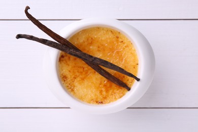 Delicious creme brulee in bowl and vanilla pods on white wooden table, top view