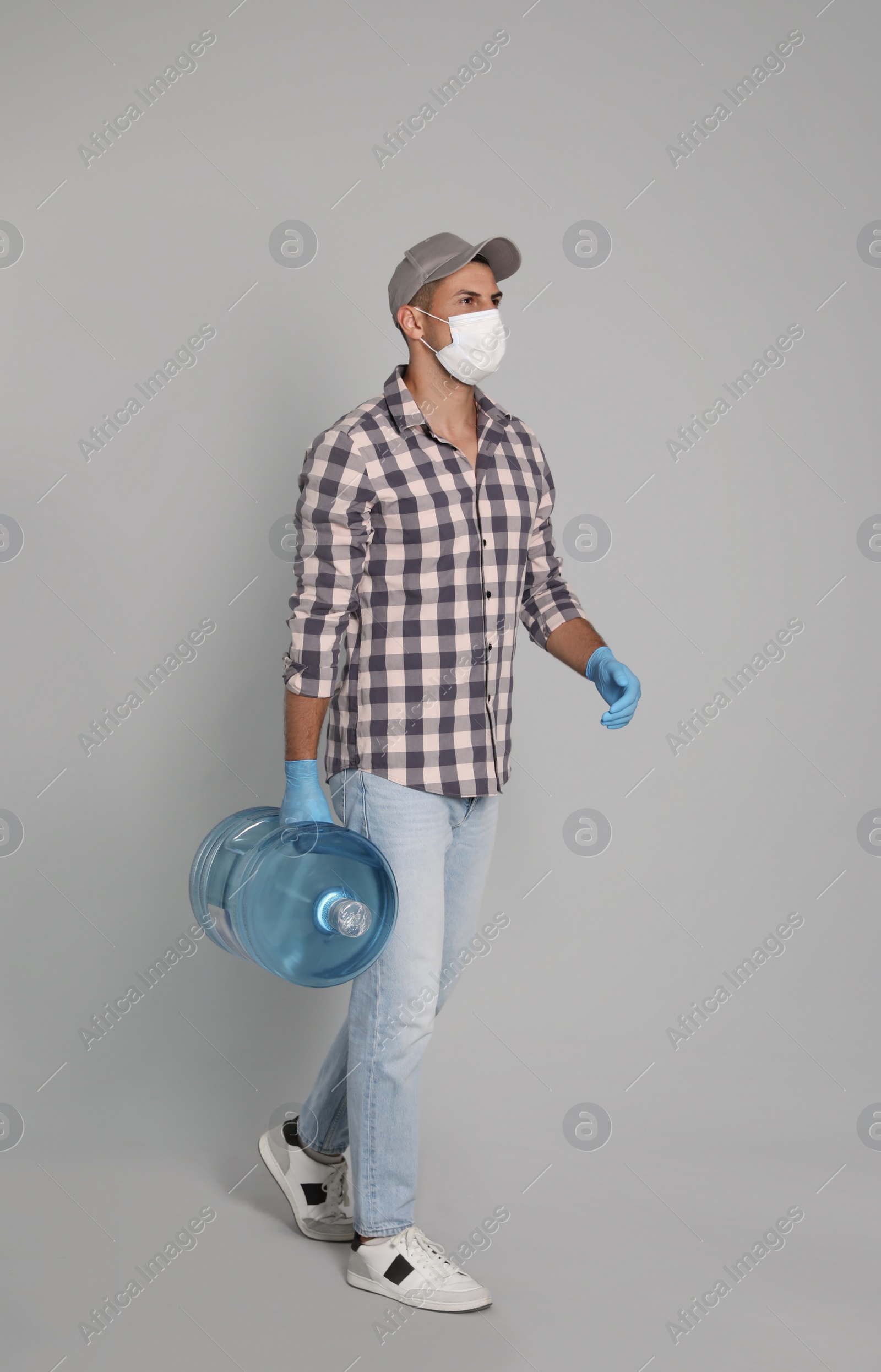 Photo of Courier in medical mask holding bottle for water cooler on light grey background. Delivery during coronavirus quarantine