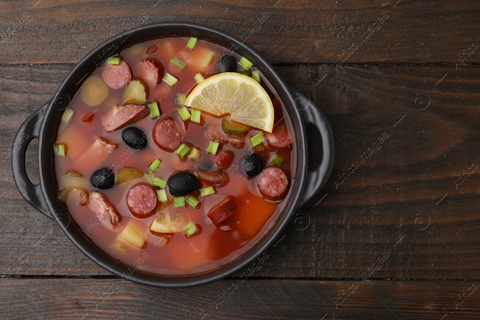 Photo of Meat solyanka soup with thin dry smoked sausages in bowl on wooden table, top view. Space for text